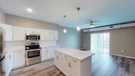 This single-floor space offers a thoughtfully designed open-concept layout that seamlessly connects the kitchen and living areas. The kitchen is a standout feature, showcasing white shaker-style cabinetry, a full suite of stainless steel appliances including a refrigerator, range, and built-in microwave, and a generous center island with a clean white countertop. Teardrop pendant lights hang above the island, complementing the recessed lighting overhead. Light wood-look flooring runs throughout the main living areas, adding warmth and continuity.

The living area extends naturally from the kitchen, bathed in natural light from a large sliding glass door fitted with vertical blinds that opens to an outdoor space. A ceiling fan provides additional comfort, and exposed ductwork adds an understated industrial accent to the design.

The bedroom is carpeted for comfort, featuring a ceiling fan, a window with blinds for natural light, and neutral taupe walls that create a calm atmosphere.

The bathroom is clean and functional, featuring a wide white vanity countertop with an undermount sink, a large mirror, and a glass-enclosed shower with tiled surround, completing this well-appointed space.