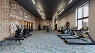 Located at 1425 Semmes Ave in Richmond, Virginia, this single-floor communal space masterfully combines industrial heritage with contemporary design. The interior is defined by soaring ceilings with exposed wooden beams and trusses, warm hardwood flooring, and prominently featured exposed brick walls that lend the space an authentic, character-rich atmosphere.

The main lounge area is generously proportioned, furnished with a mix of tufted leather sofas, barrel chairs, and accent seating arranged around coffee tables, creating multiple intimate conversation zones. A striking candelabra-style chandelier anchors the space overhead, while wall-mounted flat-screen televisions are integrated into a slatted wood feature wall. Large floor-to-ceiling windows flood the interior with natural light and offer views of the surrounding neighborhood.

A dedicated café-style area features marble-topped bistro tables, built-in banquette seating along the windows, pendant lighting, and a sleek service counter equipped with beverage amenities. A welcoming entry zone displays bold, graphic wall lettering and eclectic décor against exposed brick. An additional mailroom area presents a bold monochromatic graphic floor, resident mailboxes, and motivational wall typography, completing this thoughtfully designed, multifunctional community space.