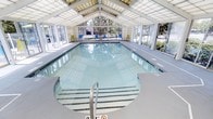This single-level indoor aquatic facility is designed to provide a premium swimming experience in a controlled, comfortable environment. At its centerpiece is a generously sized rectangular lap pool with clear, blue-tinted water, defined lane markings along the pool floor, and stainless steel entry railings for easy access.

The architecture is a standout feature of this space. A soaring vaulted ceiling fitted with large translucent skylight panels floods the interior with abundant natural light, creating a bright and inviting atmosphere throughout the day. Expansive floor-to-ceiling windows line the walls, offering views of the surrounding outdoor greenery and further enhancing the sense of openness.

Wide pool decks on all sides provide ample space for poolside seating, with bright yellow and blue chairs positioned near the far end of the pool, adding a pop of color to the otherwise clean, neutral palette of whites and grays. The overall design emphasizes functionality and comfort, making this facility well-suited for both recreational swimming and structured aquatic programs. The combination of natural light, architectural detail, and well-maintained amenities makes this an exceptional indoor aquatic space.