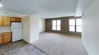 This single-floor space offers a straightforward, functional layout designed for comfortable everyday living. The living area is generously sized, featuring neutral beige carpeting that extends throughout, cream-toned walls, and a textured ceiling. Multiple windows dressed with horizontal blinds allow natural light to fill the room, while a taupe accent wall adds a subtle contrast to the otherwise neutral palette. HVAC vents are integrated into the ceiling, and the open layout provides flexibility for various furniture arrangements.

The kitchen transitions from the living area via tile flooring and is equipped with warm honey-toned wood cabinetry offering ample upper and lower storage. White appliances, including an electric coil-top range and a full-size refrigerator, complement the light countertops. An overhead range hood is positioned above the stove, and the efficient L-shaped layout maximizes workspace.

The bathroom is clean and utilitarian, featuring slate-toned tile flooring, a white tub and shower combination with chrome fixtures and a curtain rod, a toilet, and a wood-toned vanity with a white countertop. The space is bright and well-maintained, presenting a move-in ready environment.