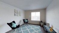This single-storey space presents a clean, functional layout designed with simplicity and comfort in mind. The living area serves as the focal point of the space, characterized by smooth, neutral-toned walls and dark hardwood-style flooring that grounds the room with a warm, contemporary feel. A large window allows natural light to fill the space, creating a bright and inviting atmosphere while offering a view of the surrounding outdoor environment.

The room is well-proportioned, providing ample space for seating and relaxation. A plush, light blue trellis-patterned area rug adds texture and a pop of colour, complementing the room's soft, neutral palette. Butterfly-style accent chairs with teal cushions are arranged thoughtfully around the space, contributing to a casual yet stylish ambiance. Wall art featuring botanical motifs adds a decorative touch to the otherwise understated walls.

The smooth, white ceiling enhances the sense of openness throughout the room. Overall, the space strikes a balance between minimalism and comfort, making it a practical and pleasant environment suitable for everyday living.