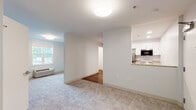 Located at 873 Harwich Rd in Brewster, Massachusetts, this single-floor residence offers a clean, modern interior designed for comfortable everyday living. The open layout connects a carpeted living area to a compact, well-equipped kitchen, creating a cohesive and functional flow throughout the space. Natural light filters in through windows, brightening the neutral white walls and light-toned carpeting that extend across the main living and bedroom areas.

The kitchen is outfitted with stainless steel appliances, including a refrigerator, electric range, and over-the-range microwave, all complemented by white shaker-style cabinetry and granite-look countertops. Wood-look flooring grounds the kitchen and bathroom areas, providing a practical contrast to the soft carpet elsewhere.

The bedroom is generously sized, with multiple doorways leading to closet space and adjoining rooms, offering a sense of separation and privacy within the single-floor plan. The bathroom is thoughtfully designed with accessibility in mind, featuring grab bars, a roll-in shower with a fold-down bench, a handheld showerhead, and a vanity with a granite-look countertop. The entry area provides a welcoming transition into the home. Overall, the space presents a bright, clean, and practical environment with modern finishes throughout.