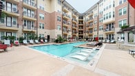 This thoughtfully designed residential unit is situated within a striking multi-story building that showcases a blend of brick and stone façades, with private balconies overlooking a beautifully appointed courtyard. The outdoor amenity space centers around a generously sized swimming pool surrounded by lounge chairs, offering a resort-like atmosphere for residents.

Inside, the unit features warm, neutral-toned walls and dark hardwood-style flooring that lend a sophisticated, cohesive aesthetic throughout. The dining area serves as a focal point, anchored by a stunning round green marble pedestal table paired with woven rattan chairs atop a textured area rug. A large framed abstract artwork adorns the wall, adding an artistic touch to the space.

A standout architectural detail is the sliding barn door, mounted on a black iron track, which adds an industrial-chic element while offering flexible room separation. Recessed lighting illuminates the interiors cleanly and efficiently. The layout connects seamlessly to adjacent rooms, visible through glass-paneled interior doors that hint at additional curated living spaces featuring gallery-style wall art. The overall ambiance is modern yet warm, combining eclectic furnishings with clean architectural lines for a lived-in yet refined feel.
