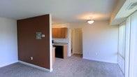 This single-floor space offers a practical and well-organized open layout that seamlessly connects the living area to the kitchen and beyond. The living area is finished with wall-to-wall carpet in a neutral tone, creating a warm and comfortable environment underfoot. A bold brown accent wall adds a touch of character and visually anchors the space, providing a distinctive contrast to the surrounding cream-colored walls and white trim.

The kitchen is efficiently designed, featuring warm honey-toned wood cabinetry that provides ample storage, complemented by granite-look countertops that add a refined touch. A black appliance suite lends a sleek, modern contrast to the cabinetry. The kitchen transitions naturally toward a hallway that leads to additional rooms, suggesting a functional and connected floor plan.

Ceiling lighting in the living area provides consistent, even illumination throughout. The clean white baseboards and neutral palette throughout create a cohesive, move-in-ready atmosphere. Overall, this space offers a straightforward and efficient layout suited to everyday living, with design elements that balance practicality with modest aesthetic appeal.