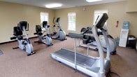 This single-floor fitness center is designed to provide a functional and comfortable environment for cardio-focused workouts. The room features warm beige walls and durable carpet flooring throughout, creating a clean and inviting atmosphere. The space is brightened by a combination of large overhead fluorescent panel lighting and multiple windows along the walls, allowing natural light to filter in and providing views of the surrounding exterior.

The fitness center is equipped with a comprehensive selection of cardio machines, including treadmills — notably the Vision T80 model — elliptical cross-trainers, and both upright and recumbent stationary bikes. The equipment is arranged to allow comfortable circulation around each machine. A wall-mounted flat-screen television is positioned for easy viewing during workouts, adding an entertainment element to training sessions.

The room features a dedicated entry door with an illuminated exit sign, and a doorway visible in one corner leads to an adjacent space with tiled flooring, suggesting a connected utility or restroom area. Climate control is provided via ceiling vents, ensuring comfort year-round. Overall, this fitness center offers a practical, well-maintained space suited for daily cardiovascular exercise routines.