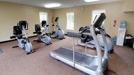 This single-floor fitness center is designed to provide a functional and comfortable environment for cardio-focused workouts. The room features warm beige walls and durable carpet flooring throughout, creating a clean and inviting atmosphere. The space is brightened by a combination of large overhead fluorescent panel lighting and multiple windows along the walls, allowing natural light to filter in and providing views of the surrounding exterior.

The fitness center is equipped with a comprehensive selection of cardio machines, including treadmills — notably the Vision T80 model — elliptical cross-trainers, and both upright and recumbent stationary bikes. The equipment is arranged to allow comfortable circulation around each machine. A wall-mounted flat-screen television is positioned for easy viewing during workouts, adding an entertainment element to training sessions.

The room features a dedicated entry door with an illuminated exit sign, and a doorway visible in one corner leads to an adjacent space with tiled flooring, suggesting a connected utility or restroom area. Climate control is provided via ceiling vents, ensuring comfort year-round. Overall, this fitness center offers a practical, well-maintained space suited for daily cardiovascular exercise routines.