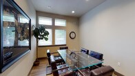 This single-story space presents a thoughtfully designed meeting or conference room that combines functionality with a polished, contemporary look. The room is anchored by a sleek glass-top table surrounded by dark leather chairs, creating a professional yet inviting atmosphere suited for meetings or collaborative work. Recessed lighting in the smooth ceiling supplements the natural light that filters in through a set of windows fitted with horizontal blinds, offering both illumination and a view of the greenery outside. The neutral-toned walls provide a clean backdrop, while wood-look flooring adds warmth and texture to the space. A wall-mounted flat-screen display on one side enhances the room's utility for presentations, and a classic round wall clock serves as a practical accent. A potted fiddle-leaf fig plant in the corner introduces a touch of nature, softening the room's modern edge. Overall, the space conveys a well-maintained and organized environment with a balance of professional design elements and comfortable furnishings.