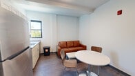 Located at 601 Clyde St in Pittsburgh's Oakland neighborhood, this one-bedroom, two-bathroom unit offers a functional and modern single-floor layout. The living and dining area features dark hardwood-style flooring, crisp white walls, and a window that brings in natural light with views of surrounding greenery. A compact kitchen with light cabinetry and a sink is integrated seamlessly into the living space, keeping the layout efficient and open.

The bedroom is a well-proportioned, carpeted room with large black-framed windows that frame tree-lined outdoor views, filling the space with natural light. A dedicated desk area within the bedroom provides a convenient workspace. An additional office room offers flexible use as a study or workspace.

Both bathrooms are finished in a clean, contemporary style with neutral tile flooring, white vanities featuring stone-look countertops, and full tub-shower combinations with built-in shelving niches. The bathrooms are bright and well-appointed, maintaining a consistent, modern aesthetic throughout.

Two hallways connect the rooms, contributing to a logical and practical flow throughout the unit. The overall design is clean and understated, with a neutral color palette and modern fixtures that create a comfortable and functional living environment suited for individuals or students in the Pittsburgh area.