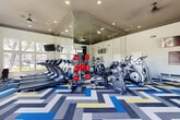 Located in Peoria, Arizona, this well-equipped fitness center offers a comprehensive workout environment designed to support a wide range of exercise routines. The space features soaring ceilings with recessed lighting that illuminate the room evenly, creating a bright and energizing atmosphere throughout.

The exercise room is outfitted with an extensive selection of equipment, including multiple treadmills, elliptical trainers, stationary bikes, and cable weight machines, along with free weights, dumbbells, medicine balls, and adjustable weight benches. A dedicated strength training area provides additional versatility for resistance-based workouts.

A full wall of floor-to-ceiling mirrors expands the sense of space and allows users to monitor their form, while large multi-pane windows along an entire wall flood the interior with natural light and frame views of the surrounding landscaped grounds and palm trees. Wall-mounted flat-screen TVs are positioned throughout for entertainment during workouts.

The bold, geometric chevron-patterned carpet in shades of blue, gray, yellow, and white adds a distinctive visual energy to the space, complementing the modern, clean aesthetic of the white walls and dark tray ceiling details. French doors provide easy access to the outdoors, further connecting the interior to the sunny Arizona surroundings and enhancing the overall sense of openness.