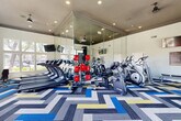 Located in Peoria, Arizona, this well-equipped fitness center offers a comprehensive workout environment designed to support a wide range of exercise routines. The space features soaring ceilings with recessed lighting that illuminate the room evenly, creating a bright and energizing atmosphere throughout.

The exercise room is outfitted with an extensive selection of equipment, including multiple treadmills, elliptical trainers, stationary bikes, and cable weight machines, along with free weights, dumbbells, medicine balls, and adjustable weight benches. A dedicated strength training area provides additional versatility for resistance-based workouts.

A full wall of floor-to-ceiling mirrors expands the sense of space and allows users to monitor their form, while large multi-pane windows along an entire wall flood the interior with natural light and frame views of the surrounding landscaped grounds and palm trees. Wall-mounted flat-screen TVs are positioned throughout for entertainment during workouts.

The bold, geometric chevron-patterned carpet in shades of blue, gray, yellow, and white adds a distinctive visual energy to the space, complementing the modern, clean aesthetic of the white walls and dark tray ceiling details. French doors provide easy access to the outdoors, further connecting the interior to the sunny Arizona surroundings and enhancing the overall sense of openness.