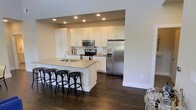 This single-floor space showcases a thoughtfully designed open-concept layout that emphasizes both functionality and modern style. The kitchen is a central highlight, outfitted with a full suite of stainless steel appliances, including a French door refrigerator with ice maker, a stainless range, and an over-the-range microwave. Crisp white shaker-style cabinetry lines the walls, offering generous storage space, while a classic subway tile backsplash adds a clean, timeless aesthetic. A spacious kitchen island with a light quartz countertop serves as both a prep area and a casual dining spot, accommodating several black round-back bar stools. Recessed lighting throughout the kitchen area ensures the space is well-lit and inviting. The rich, dark hardwood flooring flows throughout, providing a warm and elegant contrast to the bright white cabinetry and neutral wall tones. The open design creates a seamless connection between the kitchen and the adjacent living areas, making the space ideal for both everyday living and entertaining. Overall, the space presents a contemporary, move-in-ready environment with a cohesive and polished aesthetic.