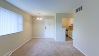 This single-floor residence offers a clean, functional layout designed for comfortable everyday living. The main living and dining area is spacious and open, finished with plush wall-to-wall carpeting that adds warmth underfoot. Neutral beige and light green walls create a calm, welcoming atmosphere throughout the space. A classic chandelier hangs above the dining area, providing a focal point and elegant touch to the room. Natural light filters in through a window along the wall, brightening the interior. The front entry door is centrally positioned and features a standard deadbolt, lending a straightforward and practical entryway. Adjacent to the living area, the kitchen is accessible through an open pass-through cutout in the wall, promoting a connected and sociable flow between spaces. The kitchen is equipped with white cabinetry, under-cabinet storage, a built-in dishwasher, and a sink positioned beneath upper cabinets. The overall design is simple and uncluttered, making the space easy to personalize. With its open plan, neutral palette, and practical kitchen layout, this residence provides a comfortable and well-organized living environment on a single level.