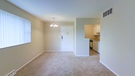 This single-floor residence offers a clean, functional layout designed for comfortable everyday living. The main living and dining area is spacious and open, finished with plush wall-to-wall carpeting that adds warmth underfoot. Neutral beige and light green walls create a calm, welcoming atmosphere throughout the space. A classic chandelier hangs above the dining area, providing a focal point and elegant touch to the room. Natural light filters in through a window along the wall, brightening the interior. The front entry door is centrally positioned and features a standard deadbolt, lending a straightforward and practical entryway. Adjacent to the living area, the kitchen is accessible through an open pass-through cutout in the wall, promoting a connected and sociable flow between spaces. The kitchen is equipped with white cabinetry, under-cabinet storage, a built-in dishwasher, and a sink positioned beneath upper cabinets. The overall design is simple and uncluttered, making the space easy to personalize. With its open plan, neutral palette, and practical kitchen layout, this residence provides a comfortable and well-organized living environment on a single level.