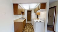 This single-story space includes a galley-style kitchen designed for efficient use of space. The kitchen features warm-toned wood cabinetry lining both walls, providing ample upper and lower storage. Light countertops offer a clean, neutral work surface, complemented by a stainless steel double-basin sink positioned along one wall. White appliances, including a coil-burner electric range with an overhead exhaust hood and a full-sized refrigerator, are integrated seamlessly into the layout. A white dishwasher is also present, adding to the kitchen's practical functionality. The flooring consists of wood-look planks in a rich, dark tone that contrasts nicely with the lighter cabinetry and countertops. Overhead, a large grid-style drop ceiling light panel provides broad, even illumination throughout the kitchen. The corridor-style layout leads toward an interior door at the far end, connecting to other areas of the space. The overall aesthetic is practical and straightforward, with a classic design that prioritizes storage and functionality.