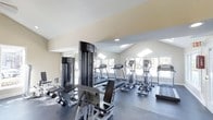 This fitness center is a thoughtfully designed, single-floor facility built to support a comprehensive workout experience. The space features vaulted ceilings that lend an open, airy feel, complemented by large windows along the walls that allow abundant natural light to fill the room throughout the day. Recessed lighting provides additional illumination, ensuring the space is well-lit at all hours. The flooring is a durable, dark-toned material well-suited for high-traffic athletic use. A full wall of mirrors enhances the sense of spaciousness while allowing users to monitor their form during exercise. The cardio section is prominently equipped with multiple treadmills and elliptical machines, arranged in a row facing the windows to take advantage of the natural views and light. Alongside the cardio equipment, the facility includes a multi-station cable strength machine and a dedicated weight bench with a selection of free weights, catering to a variety of strength training routines. The neutral, warm-toned walls contribute to a clean and focused atmosphere. Overall, the fitness center is well-organized, offering a balanced mix of cardiovascular and resistance training equipment within a bright, inviting environment.