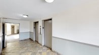 This single-floor space presents a practical and utilitarian elevator lobby area. The corridor features two side-by-side elevator doors finished in a muted gray-brown tone, each clearly numbered and equipped with standard call panels on the adjacent wall. The walls are painted in a neutral off-white above a contrasting sage green wainscoting that runs along the lower portion, creating a simple two-tone aesthetic. The flooring consists of natural stone-style tiles in warm earth tones — tan, ochre, and brown — laid in a grid pattern that adds texture to the space. Ceiling-mounted flush light fixtures provide adequate ambient illumination throughout the corridor. The overall design is straightforward and functional, prioritizing accessibility and ease of navigation. The clean lines and minimal ornamentation reflect a no-frills, practical approach to shared building infrastructure. The space is well-maintained, with a tidy and orderly appearance that serves its purpose as a transitional area connecting different parts of the building.