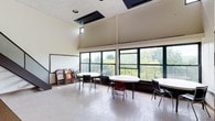 This single-floor multipurpose space is designed to accommodate a variety of uses, from meetings and classes to community gatherings. The room's most striking architectural feature is its vaulted ceiling, which rises dramatically to create a sense of openness and volume well beyond what a standard ceiling would allow. A clerestory window near the peak of the ceiling draws in additional natural light, complementing the generous illumination provided by a continuous band of large, black-framed windows along the exterior wall. These windows frame sweeping views of mature trees, bringing a refreshing connection to the outdoors into the interior. The flooring is a durable polished concrete surface, well-suited to high-traffic use. Round tables paired with folding chairs are arranged throughout the room, offering flexible seating configurations that can be easily reconfigured as needed. Additional stacked folding chairs are stored along one wall, allowing the space to scale up in capacity. A whiteboard is mounted near the entry area, supporting instructional or presentational activities. Ceiling-mounted HVAC units ensure climate control throughout the space. Overall, the room combines functional, adaptable furnishings with bright, airy architecture to create a welcoming and practical communal environment.