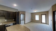This single-floor space features an open-concept design that connects the kitchen and living areas in a seamless, functional layout. The kitchen is appointed with dark wood cabinetry, including a tall pantry-style cabinet, and granite countertops with a curved edge that transitions naturally into the living space. Recessed and flush-mount ceiling lighting provides consistent illumination throughout.

The living area is finished with neutral beige carpeting and warm tan walls accented by dark brown trim around doors and baseboards, creating a cohesive and grounded color palette. A wall-mounted air conditioning unit and baseboard heating ensure year-round climate control. Two windows allow natural light to brighten the space, while a glazed exterior door offers additional light and direct access to the outside.

An open doorway leads to an adjacent room, suggesting additional private living quarters beyond the main area. The layout is straightforward and practical, with a comfortable sense of openness despite its modest footprint. Overall, the space presents a clean, move-in ready environment with functional finishes throughout.
