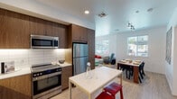 This well-designed single-floor unit showcases a thoughtful open-concept layout that seamlessly connects the kitchen, dining, and living areas. The kitchen is a standout feature, equipped with rich dark wood cabinetry extending to the ceiling, a white subway tile backsplash, white quartz countertops with under-cabinet lighting, and a full suite of stainless steel appliances including a refrigerator, range, and built-in microwave. A white kitchen island with bar seating adds both workspace and casual dining functionality. Light wood-style flooring flows throughout the main living areas, enhancing the bright, airy feel created by multiple windows dressed with clean white blinds. The living and dining areas are open and well-lit, offering a comfortable and functional layout. The unit includes multiple bedrooms, each carpeted and fitted with ceiling fans, large windows for natural light, and ample space. The bathrooms feature wood-tone vanities with white quartz countertops, tiled tub-shower combinations, and light wood-style flooring. One bathroom includes a double-sink vanity and open shelving for additional storage. Throughout, the space maintains a cohesive modern aesthetic with clean white walls and contemporary fixtures.