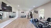 Located at 4727 Grant Line Rd in New Albany, Indiana, this single-floor fitness facility offers a well-appointed and thoughtfully designed workout environment. The main exercise area is generously proportioned, featuring light wood-look flooring throughout that lends a clean, modern aesthetic. Recessed lighting illuminates the space evenly, creating a bright and energizing atmosphere.

The primary workout floor is equipped with a multi-station cable and weight machine, providing a comprehensive strength training setup. A dedicated free weights zone includes a flat weight bench, a full rack of dumbbells, and additional weight plates, all positioned atop protective rubber floor mats. Full-length mirrors line one wall, allowing users to monitor form during exercise. Motivational artwork and signage are displayed throughout, reinforcing an active atmosphere.

A cardio section houses two elliptical machines, positioned near a window that allows natural light into the space. A wall-mounted flat-screen television provides entertainment during workouts. An adjacent side room offers additional storage and workout space, complete with a double closet, a flat bench, and a dumbbell rack.

A carpeted staircase with white railings and newel posts connects to an upper level, accompanied by a wall-mounted water fountain nearby. The facility's neutral color palette, ample floor space, and organized layout make it a functional and inviting space for a full range of fitness activities.