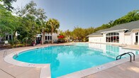 Located at 3301 Glenn McConnell Pkwy in Charleston, South Carolina, this outdoor amenity area showcases a generously sized resort-style swimming pool as its centerpiece. The crystal-clear blue pool is flanked by a wide concrete sun deck outfitted with lounge chairs and red umbrella-shaded seating, providing ample space for relaxation under the South Carolina sun. Towering palm trees and mature hardwoods frame the perimeter, lending a lush, tropical ambiance that reflects the character of the Lowcountry. A charming single-story clubhouse building anchors one end of the pool area, featuring arched windows, a peaked gazebo-style roofline with a cupola, and an adjacent outdoor patio with additional dining seating. A stone outdoor fireplace is also visible near the patio, extending the usability of the outdoor space. A secondary utility or support structure with white siding and traditional windows is positioned nearby. Iron fencing defines the boundary of the pool and patio areas, while mature trees provide natural shade and privacy. A separate shaded patio area with metal table-and-chair seating nestled among palm trees offers a quieter retreat. The overall setting is well-maintained and thoughtfully landscaped, creating an inviting outdoor gathering environment.