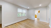 This single-floor residence offers a clean, functional layout with a bright and neutral aesthetic throughout. The living areas are finished with light hardwood flooring that extends through the main spaces, complemented by crisp white walls and recessed lighting that create an open, airy feel. Natural light flows in through multiple windows fitted with white horizontal blinds, offering views of surrounding greenery and the neighboring streetscape. Baseboard heating is present throughout, ensuring consistent comfort year-round. The kitchen is equipped with warm honey-toned wood cabinetry, a mosaic tile backsplash in earthy tones, dark countertops, a gas range, and a full-size refrigerator, with a window above the sink providing additional natural light. The bedrooms feature plush carpeting and their own windows with neighborhood views, while one additional room with hardwood flooring includes a closet and access to the outdoors. A hallway connects the rooms, transitioning between hardwood and carpeted areas. The bathroom showcases large-format beige ceramic tile surrounding the bathtub, accented by a decorative mosaic border strip in warm brown and amber tones. Overall, the space presents a well-kept, move-in ready environment with practical finishes and a welcoming atmosphere.
