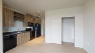 This single-floor unit presents a clean, contemporary design throughout its open-concept layout. Light-toned luxury vinyl plank flooring runs continuously across the living and sleeping areas, contributing to a bright, cohesive aesthetic.

The kitchen is well-appointed with medium-toned wood-grain cabinetry, dark laminate countertops, and a full suite of black appliances, including an electric range, over-the-range microwave, refrigerator, and dishwasher. A stainless steel sink and track lighting overhead complete the functional workspace.

The living area flows naturally from the kitchen and benefits from a large multi-pane window that frames views of the surrounding cityscape, allowing natural light to fill the space. A dedicated wet bar or secondary counter area with upper cabinetry and a sink adds versatility to this zone.

The bedroom is generously proportioned, featuring neutral walls and a sliding double-panel closet door that opens to a built-in shelving system for organized storage.

The bathroom maintains the same design language found throughout the unit, with wood-grain cabinetry, a dark countertop vanity, and light flooring. A full tub and shower combination, toilet, and towel bar complete the space efficiently.
