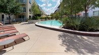 This outdoor amenity space is designed to provide a resort-like experience within a residential setting. At its centerpiece is a generously sized, curved swimming pool with clear blue water, surrounded by a spacious concrete pool deck that accommodates a collection of red lounge chairs and umbrella-shaded seating areas — ideal for sunbathing and socializing. Mature trees, ornamental grasses, and native plantings are thoughtfully integrated throughout the landscape, lending the area a lush, verdant character.

Adjacent to the pool deck is a covered pergola structure supported by substantial wood timber posts, offering a shaded transition zone between the interior building and the outdoor space. Beneath the pergola, metal outdoor dining tables and chairs provide a comfortable setting for al fresco gatherings. Floor-to-ceiling glass walls and sliding glass doors connect the interior clubroom or lounge seamlessly to the pergola and pool area, allowing natural light to flood the interior while maintaining a visual and physical connection to the outdoors. The surrounding multi-story residential buildings frame the courtyard, creating an intimate and private atmosphere. Overall, the space balances modern architectural design with inviting, functional outdoor living areas.