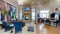 This single-floor space is designed with both comfort and functionality in mind, offering a well-appointed interior that seamlessly blends a lounge area with a kitchen and dining space. The focal point of the room is a striking floor-to-ceiling stacked stone fireplace, above which a wall-mounted flat-screen television is positioned for easy viewing. The fireplace is flanked by tall windows dressed with neutral curtains, allowing generous natural light to fill the space and highlight the warm hardwood flooring throughout.

The lounge area is furnished with plush blue accent chairs, a round coffee table, and a cushioned bench ottoman, all anchored by a light area rug that adds texture and definition to the seating zone. A geometric, colorful art piece on one wall contributes a modern and vibrant aesthetic. An elegant chandelier overhead adds a refined touch to the overall design.

To the right, the kitchen and dining area features dark cabinetry, stainless steel appliances, and a high-top bar-style table with chairs, making it ideal for casual dining or socializing. The open layout connecting both areas promotes a sense of spaciousness and encourages interaction, while crown molding and vaulted ceiling details add an architectural sophistication to the entire space.