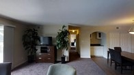 This single-floor suite offers a thoughtfully arranged living space designed for both comfort and functionality. The main living area serves as the heart of the suite, featuring warm carpeted flooring, a media console with a flat-screen television, and lush indoor plants that add a natural, refreshing touch to the interior ambiance. A plush seating area invites relaxation, making the space feel welcoming and well-appointed. A hallway connects the living area to a private bedroom, which is visible in the background and provides a quiet retreat with its own warm lighting. An elegant arched doorway leads to a kitchenette area, which is outfitted with white cabinetry and a dark countertop, offering convenient in-suite amenities. The transition between carpeted and hard flooring zones subtly defines the different functional areas within the suite. Textured ceilings and neutral wall tones contribute to the overall cozy and understated aesthetic throughout. The layout flows naturally from the living space to the sleeping and utility areas, creating a cohesive and practical environment suited for extended stays or comfortable everyday living.