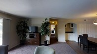 This single-floor suite offers a thoughtfully arranged living space designed for both comfort and functionality. The main living area serves as the heart of the suite, featuring warm carpeted flooring, a media console with a flat-screen television, and lush indoor plants that add a natural, refreshing touch to the interior ambiance. A plush seating area invites relaxation, making the space feel welcoming and well-appointed. A hallway connects the living area to a private bedroom, which is visible in the background and provides a quiet retreat with its own warm lighting. An elegant arched doorway leads to a kitchenette area, which is outfitted with white cabinetry and a dark countertop, offering convenient in-suite amenities. The transition between carpeted and hard flooring zones subtly defines the different functional areas within the suite. Textured ceilings and neutral wall tones contribute to the overall cozy and understated aesthetic throughout. The layout flows naturally from the living space to the sleeping and utility areas, creating a cohesive and practical environment suited for extended stays or comfortable everyday living.