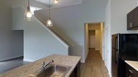 This two-storey space is characterized by its clean, modern design and thoughtful layout. The kitchen is a standout feature, equipped with a double stainless steel sink set into a granite-look countertop with dark cabinetry below. A pendant light with an exposed bulb hangs above, adding a contemporary touch. The soaring ceiling height in this area creates an impressive sense of openness, drawing the eye upward toward a skylight that allows natural light to filter in.

A staircase with a white-painted railing rises prominently alongside the kitchen, connecting the two floors while contributing to the architectural character of the space. The walls are finished in a soft grey tone throughout, providing a neutral and cohesive backdrop. Warm hardwood flooring runs through the main level, extending down a hallway that leads to additional rooms. White-trimmed doorways line the corridor, offering glimpses of further spaces beyond. The overall ambiance is bright, airy, and well-appointed, with a balance of functional design and modern aesthetic details evident throughout both levels.