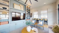 Located in Hutto, TX, this single-floor community space offers a well-appointed, open-plan layout that seamlessly integrates a living room, dining area, and full kitchen. The living room is anchored by a striking floor-to-ceiling built-in entertainment center finished in a soft powder blue, complete with open shelving, cabinet storage, and shiplap detailing. Comfortable seating arrangements and a large ceiling fan contribute to a relaxed yet polished atmosphere, while multiple large windows fitted with roller shades allow abundant natural light and neighborhood views. The dining area features warm wide-plank hardwood flooring, marble-top round tables, and cane-back chairs, all illuminated by oversized woven rattan pendant lights that lend a coastal, casual-chic aesthetic. Built-in banquette seating along one wall adds functional charm. The kitchen is outfitted with crisp white cabinetry, grey subway tile backsplash, quartz countertops, open wood shelving, stainless steel appliances including a double wall oven and French door refrigerator, and a central island with bar seating. Gold-toned pendant fixtures add a refined touch. The cohesive design palette of blues, whites, and natural wood tones creates a welcoming, community-focused environment throughout.