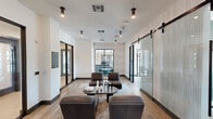 Located in Phoenix, Arizona, this thoughtfully designed amenity space offers a seamless blend of comfort and functionality across a single floor. The layout includes a central living lounge and two dedicated conference rooms, making it well-suited for both relaxed gatherings and professional meetings.

The main lounge area sets the tone with warm wood-look flooring, textured accent wall panels, and a curated seating arrangement featuring deep leather chairs and ottomans centered around a sleek coffee table. Pendant and track lighting provide a polished ambiance, while a built-in wet bar with teal cabinetry adds a vibrant design accent. Sliding barn-style doors with frosted ribbed glass panels elegantly divide the lounge from the conference rooms, maintaining visual openness while offering privacy on demand.

The first conference room is a larger space anchored by a substantial wood oval table surrounded by upholstered task chairs, built-in shelving, and a window overlooking the surrounding Phoenix streetscape. A striking oversized dome pendant light with a gold interior adds warmth to the room.

The second, more intimate conference room features a round dark table, a wall-mounted electric fireplace framed in a deep gray accent wall, and a matching oversized pendant light, creating a focused yet inviting setting. Natural light filters in through a large window, offering views of the neighborhood beyond.