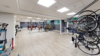 Located at 1704 Shelburne Rd in South Burlington, Vermont, this single-floor amenity space offers a generous and versatile layout designed to serve the recreational and practical needs of its users. The space is finished with light wood-look luxury vinyl plank flooring throughout, providing a clean and modern aesthetic that ties the various zones together seamlessly.

A standout feature is the expansive bicycle storage area, which occupies a substantial portion of the floor plan. Multiple freestanding metal bike racks accommodate a large number of bicycles both vertically and horizontally, making this an exceptional amenity for cycling enthusiasts. The walls in this section are painted in a bold orange accent color, adding energy and visual interest to the room.

The lounge and recreation zone is defined by a full-size pool table with a vibrant red felt surface, complemented by a plush orange sofa and a foosball table — creating a welcoming social hub. Recessed lighting and flush-mounted LED panel lights in a drop tile ceiling ensure the entire space is evenly and brightly illuminated. Small windows along one wall allow natural light to filter in.

Additional features include what appears to be a utility or kitchenette area with cabinetry, as well as ample open floor space that allows for flexible use. The overall ambiance is modern, functional, and community-focused.