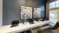 Located at Rocky Ridge Blvd in Douglasville, Georgia, this single-floor community or leasing office presents a polished, contemporary interior designed to impress and inform visitors. The space is thoughtfully appointed throughout, blending professional functionality with upscale residential aesthetics.

The business center area features a long, marble-topped workstation accommodating multiple workstations, complemented by sleek leather task chairs and lush greenery. A pair of striking abstract artworks mounted on deep slate-blue walls adds a sophisticated artistic touch to the workspace.

Adjacent lounge and seating areas are furnished with plush leather armchairs, geometric-patterned area rugs in black and gold tones, and warm accent lighting from floor lamps. Crown molding and tall ceilings lend an air of elegance, while large windows dressed in floor-length drapes flood the space with natural light and offer views of the surrounding landscape.

A display area visible from the main lounge showcases material and finish samples, catering to prospective residents exploring their options. The open sightlines between rooms create a seamless flow, reinforced by consistent warm-gray and muted teal wall tones throughout. Pendant and chandelier lighting further enhance the welcoming, upscale ambiance of this well-curated community space.