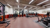 Located at 1525 N American St in Philadelphia's vibrant 19122 corridor, this fitness center offers a comprehensive exercise environment across a single, open floor. The space is defined by its industrial-inspired aesthetic, featuring exposed concrete ceilings with visible ductwork and structural beams, all illuminated by rows of bright linear LED lighting that ensure excellent visibility throughout.

The main workout area is finished with durable red rubber flooring and features an extensive selection of cardio equipment, including treadmills, stair climbers, stationary bikes, and rowing machines. A dedicated strength training zone houses a cable machine, power rack, and a full dumbbell rack with adjustable benches. Floor-to-ceiling mirrors line multiple walls, enhancing the sense of space and allowing users to monitor their form. A bold red wainscoting detail adds a striking visual contrast to the white upper walls.

Adjacent to the main floor is a separate studio room, finished with warm cork-style hardwood flooring and its own mirrored wall. This versatile space is equipped with free weights, balance trainers, stability balls, and resistance tools, making it ideal for functional training, stretching, or group classes. The combination of dedicated zones, quality equipment, and a clean industrial design makes this a fully functional and well-appointed fitness facility.