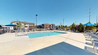 This single-story community clubhouse is designed to offer residents a well-rounded amenity experience, blending indoor and outdoor spaces seamlessly. The building's exterior showcases a craftsman-inspired aesthetic, featuring a gabled roofline, a covered pergola with warm wood lattice, and brick accent details that add texture and character.

The outdoor area is a central highlight, anchored by a generously sized swimming pool surrounded by a broad concrete deck lined with lounge chairs and shaded dining areas with blue umbrellas. Adjacent to the pool, a pergola-covered patio serves as an outdoor living and entertaining hub, complete with a stone-clad built-in grill station, comfortable cushioned seating, hanging planters, and open views of the surrounding green space.

Entry into the building is through bold black double French doors, which frame views of the pool beyond and allow natural light to flood the tiled entryway. The interior corridors feature light-toned tile flooring, neutral walls, and elevated ceilings that contribute to an airy, welcoming atmosphere. Practical touches such as seating benches in the hallway and clearly marked exits reflect the facility's functional design. The layout flows from the entry toward additional common areas with large windows, maintaining a bright and open ambiance throughout.