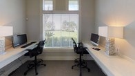 This single-floor workspace is thoughtfully designed to maximize functionality within a compact footprint. The main working area features multiple built-in, floating desks finished in white, arranged along the perimeter walls to make efficient use of the available space. Each workstation is equipped with ergonomic task seating, and the layout comfortably accommodates several users simultaneously. A large window fitted with horizontal blinds anchors one wall of the primary workspace, allowing ample natural light to fill the room while offering views of surrounding greenery. Clerestory windows positioned above the main window further enhance the brightness of the space. The neutral gray carpet throughout the workspace contributes to a calm, professional atmosphere. Dedicated alcove workstations provide additional private areas for individual tasks, each outfitted with its own built-in desk, task lamp, and storage. One alcove accommodates printing and computing equipment, while another features decorative accents including a sunburst mirror and dark cabinetry below. A transitional area with light stone tile flooring connects the workspace to adjoining spaces, maintaining a clean and cohesive aesthetic throughout. The overall design prioritizes practicality and a distraction-free environment.