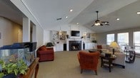 This single-floor space is defined by its vaulted ceiling, which lends a sense of openness and grandeur to the main living area. A ceiling fan with an integrated light fixture is centrally positioned, ensuring both comfort and adequate illumination throughout the room. The focal point of the living area is a classic fireplace framed by a white mantel, flanked on both sides by built-in shelving units and cabinetry. These built-ins provide generous display and storage space, adding both functionality and a polished, custom look to the room. The walls and trim are finished in crisp white, creating a clean and bright aesthetic that complements the neutral carpeted flooring. Recessed lighting is thoughtfully installed throughout the ceiling, enhancing the ambient light alongside the natural light that filters in through the windows. The living area is furnished with warm, rust-toned upholstered seating, including a sofa and accent chairs, which add a welcoming richness to the overall color palette. A wall-mounted flat-screen television sits above the fireplace, integrating modern entertainment seamlessly into the traditional design. The overall ambiance is comfortable and well-appointed, striking a balance between classic architectural details and practical, livable design.