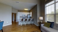 This single-floor residence offers a thoughtfully designed open-concept layout that seamlessly connects the living and kitchen areas. The kitchen is a standout feature, outfitted with crisp white cabinetry, stainless steel appliances including a refrigerator, range, and microwave, and a decorative mosaic tile backsplash that adds a touch of texture and character. A peninsula-style breakfast bar extends from the kitchen counter, accompanied by sleek metallic bar stools, creating an inviting space for casual dining or conversation. Warm hardwood flooring flows throughout the main living areas, lending a sense of continuity and comfort. The neutral taupe wall tones complement the light cabinetry and flooring, creating a cohesive and calming aesthetic. The living area is accented by natural light filtering in through windows, enhancing the open, airy feel of the space. A cozy seating area with a upholstered sofa and accent side table with a lamp adds warmth to the room. The layout is practical and well-proportioned, with additional rooms accessible via a hallway, suggesting dedicated private spaces beyond the main open-plan area.