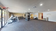 This single-floor fitness center is organized into several distinct zones, each tailored to support a variety of workout routines. The main exercise floor is generously sized and finished with durable carpet tile, housing an impressive array of equipment including treadmills, ellipticals, a multi-function cable machine, a rowing machine, and a full dumbbell rack with weight benches. A wall-mounted flat-screen television provides entertainment, while recessed lighting and crown molding lend a polished feel to the space. An adjoining dedicated spin room features warm hardwood flooring and a lineup of stationary cycling bikes, with framed workout reference charts — including band, yoga, and general exercise guides — displayed along the wall for user convenience. A separate studio area with hardwood floors, a wall-mounted TV, overhead lighting, and an interior window offers a versatile space for stretching, yoga, or small-group training, complete with a stability ball and storage cabinet. The facility also includes a spacious package locker room equipped with a large bank of smart lockers, a community bulletin board, and a sliding glass door leading to an outdoor area, adding both functionality and convenience to the overall space.