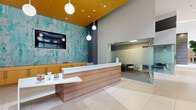 Located at 700 SW 78th Ave in Plantation, Florida, this single-floor commercial lobby space offers a sophisticated and well-appointed environment designed to impress and accommodate. The reception area serves as the focal point upon entry, featuring a sleek wood-paneled desk with a crisp white countertop, complemented by globe pendant lights and a vibrant abstract teal mural that adds an artistic energy to the space. Adjacent to the reception desk, a frosted glass-enclosed conference or meeting room provides a semi-private area for consultations. The main corridor extends generously, finished in large-format light ceramic tile flooring that runs throughout, reinforcing a clean and modern aesthetic. Soaring ceilings with warm golden wood paneling create a grand sense of height and openness. A dedicated lounge area features a white sofa, teal accent chairs, and a mirrored wall display, providing a comfortable and stylish seating arrangement. A café station, branded "The Bean Counter," is outfitted with pendant cluster lighting in gold geometric fixtures and bistro-style seating. Floor-to-ceiling windows along the perimeter flood the space with abundant natural light and frame lush palm tree-lined views of the surrounding Plantation streetscape, enhancing the overall inviting ambiance of this well-designed commercial interior.