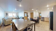 Located at 3277 Birch St in Eau Claire, Wisconsin, this single-floor community space offers a thoughtfully designed open-concept layout that seamlessly connects the kitchen, dining area, and living room. Light hardwood-style flooring flows throughout, creating a cohesive and welcoming atmosphere.

The kitchen is equipped with light maple cabinetry, white countertops, a stainless steel dishwasher, refrigerator, microwave, and a double-basin sink — providing a fully functional space for meal preparation. Two dining tables with coordinating chairs accommodate gatherings of various sizes, while a wall-mounted flat-screen TV adds a convenient entertainment element to the dining area. A large framed artwork piece adorns the wall, contributing to the space's warm, curated aesthetic.

The living area is furnished with comfortable upholstered armchairs and a sofa arranged around a central coffee table, creating an inviting setting for relaxation and conversation. A large window allows natural light to fill the room and offers views of the surrounding neighborhood. Decorative wall mirrors flank the window, enhancing the sense of openness. Recessed lighting throughout ensures the space is well-lit at all hours. The space is accessible via a key-card entry door, reflecting a secure and modern design sensibility.