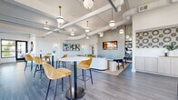 Located at 6050 Roswell Rd in Sandy Springs, GA, this single-floor combined dining and living space offers a thoughtfully designed, open-concept layout that seamlessly blends comfort with contemporary style. The interior is anchored by high ceilings with exposed ductwork and structural beams, lending an industrial-chic character balanced by a bright, white palette throughout.

The living area features a curved sectional sofa, plush sage green barrel chairs, and a distinctive organic-shaped coffee table arranged atop a woven area rug. A floor-to-ceiling textured accent wall frames a mounted flat-screen display, serving as a striking focal point. Globe pendant lights suspended from the ceiling add warmth and visual interest across the space.

The dining zone offers multiple seating configurations, including high-top bistro tables with velvet upholstered bar stools and a long communal table with modern black chairs. Large industrial-framed windows flood this area with abundant natural light and provide open views of the surrounding landscape. A roll-up glass door connects directly to an outdoor terrace, enhancing the sense of openness.

Wide-plank hardwood-style flooring runs throughout, tying the two areas together cohesively. Curated abstract artwork on the walls adds a polished, gallery-like finish to the overall ambiance.