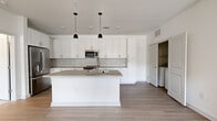This single-story residence offers a thoughtfully designed layout with two bedrooms, two bathrooms, and a connected living and kitchen area. The open-concept kitchen serves as a central highlight, featuring bright white shaker cabinetry extending to the ceiling, a hexagonal tile backsplash, stainless steel appliances including a french door refrigerator and built-in microwave, and a large center island with a quartz-style countertop and dual pendant lighting overhead. Light wood-tone flooring runs throughout the main living areas, lending warmth and continuity to the space.

The living area flows naturally from the kitchen, connected by a light-filled hallway. Both bedrooms are carpeted for comfort; the primary bedroom includes a ceiling fan and direct access to an en-suite bathroom, which features a dual-sink vanity with white cabinetry, quartz countertops, and a soaking tub with a marble-patterned tile surround. Each bedroom is served by a dedicated walk-in closet equipped with multi-tier wire shelving systems. A second full bathroom mirrors the refined finishes found throughout. An in-unit laundry closet with a full-size washer and dryer adds everyday convenience to this clean, modern residence.