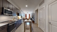 Located at 1449 S Church St in Charlotte's vibrant South End neighborhood, this single-floor residence showcases a thoughtfully designed modern interior with light wood-style flooring and a neutral color palette throughout. The galley-style kitchen is a standout feature, equipped with rich dark wood cabinetry, stainless steel GE appliances including a gas range, microwave, refrigerator, and dishwasher, complemented by quartz countertops and a geometric patterned tile backsplash. A built-in island provides additional prep and dining space, and the kitchen flows seamlessly into the open living and dining area. The living room is bathed in natural light from large windows dressed with warm, rust-colored curtains, and features a soothing slate-blue accent wall that adds depth and character to the space. The bedroom continues the warm, contemporary aesthetic with a deep teal accent wall, large windows offering outdoor views, and ample room for a full bedroom suite. The primary bathroom is generously proportioned, offering a double-sink dark wood vanity with quartz counters, a tiled soaking area, and direct access to a walk-in closet with built-in shelving. Recessed lighting and ceiling fans contribute to a comfortable and well-lit living environment throughout.