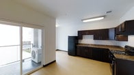 This single-floor space presents an open-concept layout that seamlessly connects the kitchen and living areas. The kitchen is outfitted with dark espresso cabinetry, granite-style countertops, a black refrigerator, and a dishwasher, all complemented by a stainless steel sink positioned beneath upper cabinets for efficient storage. A flush-mounted fluorescent light fixture provides direct illumination over the kitchen workspace. The flooring throughout is light-toned laminate, lending a clean, contemporary feel to the interior. Walls are finished in crisp white, enhancing the sense of openness. A sliding glass door connects the main living area to a private outdoor deck or balcony, where an LG wall-mounted air conditioning unit is installed, indicating a dedicated climate control system for the space. Natural light filters in through the sliding door, brightening the interior. The overall design is functional and modern, with a neutral palette that provides a versatile backdrop for a range of furnishing styles.