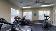 Located at 105 Doleac Drive in Hattiesburg, Mississippi, this single-floor fitness center is a dedicated amenity space reserved exclusively for residents. The room is designed with functionality and comfort in mind, offering a bright and welcoming environment for exercise. Large windows fitted with horizontal blinds line the walls, allowing generous natural light to fill the space while providing views of the surrounding exterior. A ceiling fan overhead works in tandem with recessed fluorescent lighting to maintain a comfortable atmosphere during workouts. The flooring features durable, dark rubberized material suited for fitness use, and a protective wainscoting panel runs along the lower portion of the walls for added durability. The facility is equipped with a selection of cardio machines, including multiple treadmills and an elliptical cross-trainer, catering to a variety of workout preferences. The neutral, light-toned walls contribute to an open and airy feel throughout the room. Signage clearly designates the space for resident use only, reinforcing its role as a private community amenity. Overall, the fitness center offers a clean, well-lit, and practical environment for residents to maintain an active lifestyle.