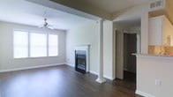 This single-floor space presents a well-appointed open layout anchored by rich, dark hardwood flooring that extends throughout the main living area. A standout architectural detail is the decorative white column with a square base and rounded capital, which adds a classic, traditional character to the interior. The focal point of the living area is a sleek fireplace framed by a crisp white mantel and set against a dark surround, providing both a functional heating element and a visual centerpiece. Natural light filters into the space through double windows dressed with horizontal blinds, brightening the neutral, warm-toned walls. A ceiling fan overhead adds practicality and visual interest to the living area. The high wall lines and clean architectural details contribute to an open, airy ambiance. An open doorway visible toward the rear of the space leads to what appears to be an additional room or storage area, suggesting a thoughtful and functional floor plan. Overall, the space balances traditional design elements with a neutral palette, creating a versatile and inviting interior.